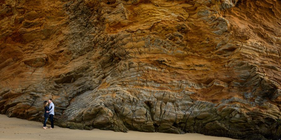 Couple small against the unique red rock cliff wall - Panther Beach Santa Cruz and Quail Hollow Felton engagement photography - Sara and Scott - photos by Bay Area wedding photographer Chris Schmauch